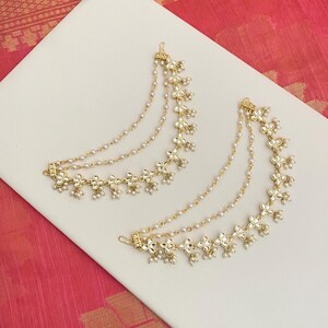 May include: Two gold-toned ear cuffs with floral and pearl accents. Each cuff features a series of delicate flowers and dangling pearls, connected by multiple strands of small pearls. The jewelry is displayed on a white surface.