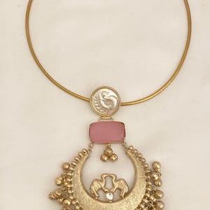 May include: Gold-toned necklace featuring a rigid collar. The pendant includes a pink rectangular stone, a circular element with a design, and a crescent-shaped piece with elephant figures and dangling beads. A small, clear gem is also present.