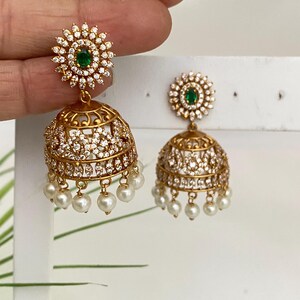 May include: Gold-toned earrings with a floral design and dangling pearls. Each earring features a cluster of clear stones around a central green stone. The lower portion is a bell shape with intricate detailing and small hanging pearls.