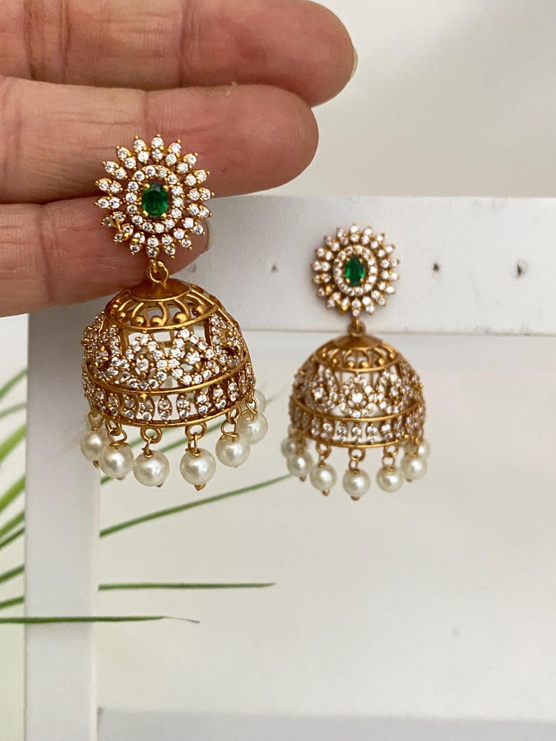 May include: Gold-toned earrings with a floral design and dangling pearls. Each earring features a cluster of clear stones around a central green stone. The lower portion is a bell shape with intricate detailing and small hanging pearls.