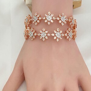 May include: Two rose gold bracelets with a floral design. Each flower has a square center with clear, sparkling stones and surrounding petals. The bracelets are worn on a person's wrist.