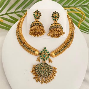 May include: A gold-toned necklace and matching pair of jhumka earrings featuring intricate metalwork and embedded green stones. The necklace has a central pendant with a circular design, while the bell-shaped earrings are adorned with small hanging gold beads.