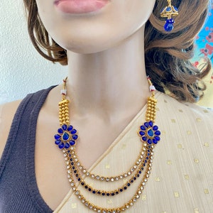 Puede incluir: Un collar de oro con tres hileras de piedras transparentes y azules. El collar tiene dos detalles de flores azules con una piedra en forma de l&aacute;grima en el centro. El collar se combina con pendientes azules a juego.