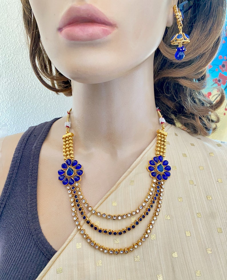 Puede incluir: Un collar de oro con tres hileras de piedras transparentes y azules. El collar tiene dos detalles de flores azules con una piedra en forma de l&aacute;grima en el centro. El collar se combina con pendientes azules a juego.