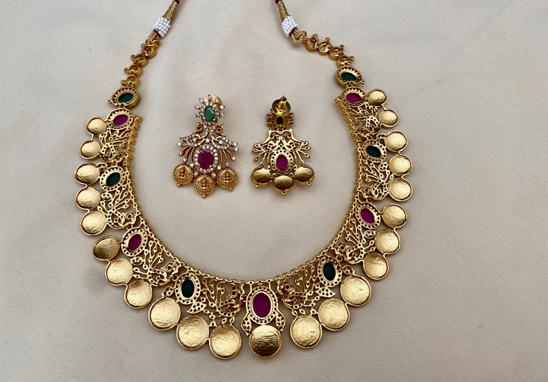 May include: A gold necklace with a coin design and pink and green gemstones. Matching earrings with pink and green gemstones.