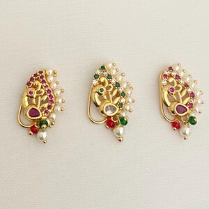 May include: Three gold-colored nose rings with a peacock design. Each ring features a central gemstone, surrounded by smaller gems and pearls. The rings have red, green, and white accents.