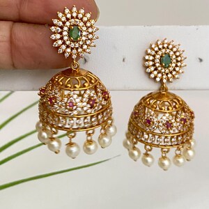May include: Gold-toned, ornate earrings with a floral design. Each earring features a central green stone surrounded by clear crystals. The lower portion is a bell shape, adorned with red stones and dangling pearls. The earrings are approximately 1.5 inches in length.