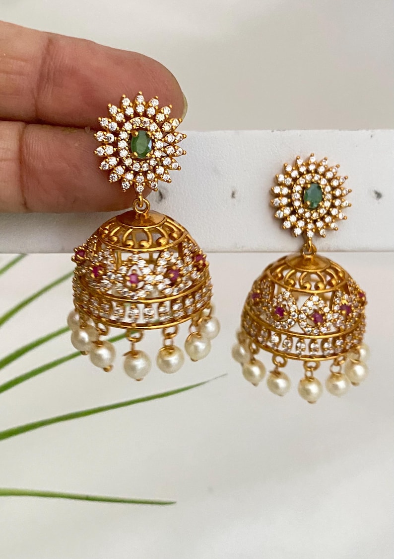 May include: Gold-toned, ornate earrings with a floral design. Each earring features a central green stone surrounded by clear crystals. The lower portion is a bell shape, adorned with red stones and dangling pearls. The earrings are approximately 1.5 inches in length.