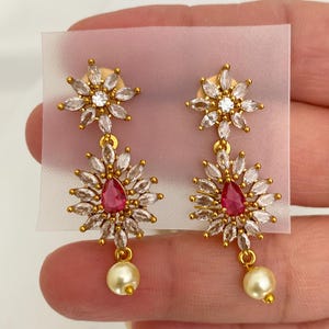 May include: Gold-toned earrings with a floral design. Each earring features a top cluster of clear crystals, a central red teardrop-shaped stone, and a dangling pearl. The earrings are set against a neutral background.