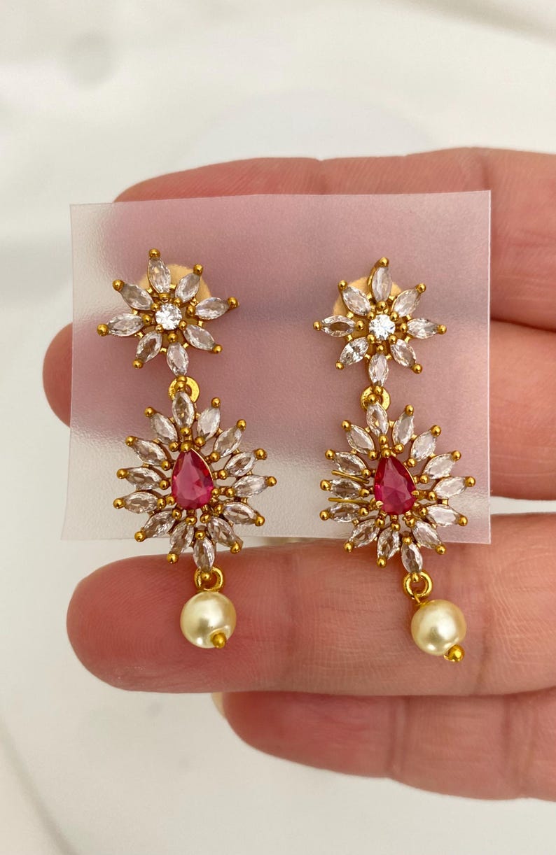 May include: Gold-toned earrings with a floral design. Each earring features a top cluster of clear crystals, a central red teardrop-shaped stone, and a dangling pearl. The earrings are set against a neutral background.