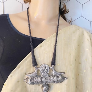 May include: A silver pendant necklace with a detailed design of two birds flanking a central figure. The pendant is suspended from a black cord.