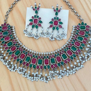 May include: A silver necklace and earrings set with red and green gemstones and white pearl accents. The necklace features a layered design with multiple rows of pearls and gemstones.
