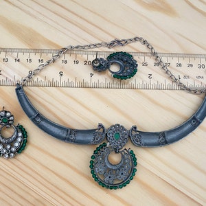 May include: A silver necklace and earrings set with green and white stones. The necklace is a crescent shape with a pendant in the center. The pendant is a circle with a crescent moon design and green stones around the edge. The earrings are also crescent shaped with green stones around the edge.