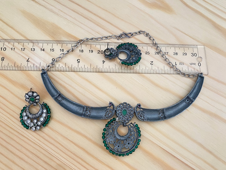 May include: A silver necklace and earrings set with green and white stones. The necklace is a crescent shape with a pendant in the center. The pendant is a circle with a crescent moon design and green stones around the edge. The earrings are also crescent shaped with green stones around the edge.