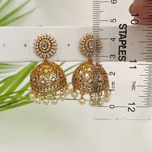May include: Gold-toned jhumka earrings with a floral design at the top and a bell-shaped bottom. The earrings are adorned with clear crystals and small, dangling pearls. The earrings measure approximately 1 inch in length.
