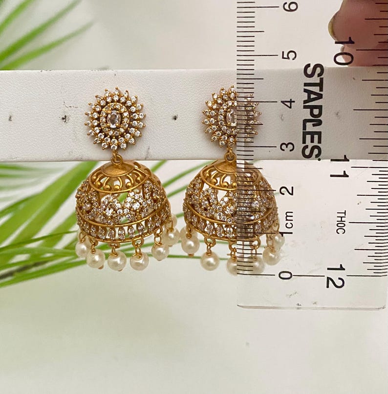 May include: Gold-toned jhumka earrings with a floral design at the top and a bell-shaped bottom. The earrings are adorned with clear crystals and small, dangling pearls. The earrings measure approximately 1 inch in length.