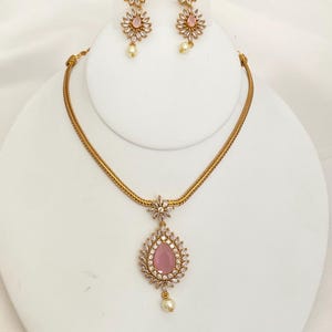 May include: A gold-toned jewelry set featuring a necklace and matching earrings. The necklace has a chain and a teardrop pendant with a pink center stone surrounded by clear crystals. The earrings have a floral design with pink stones and dangling pearls.