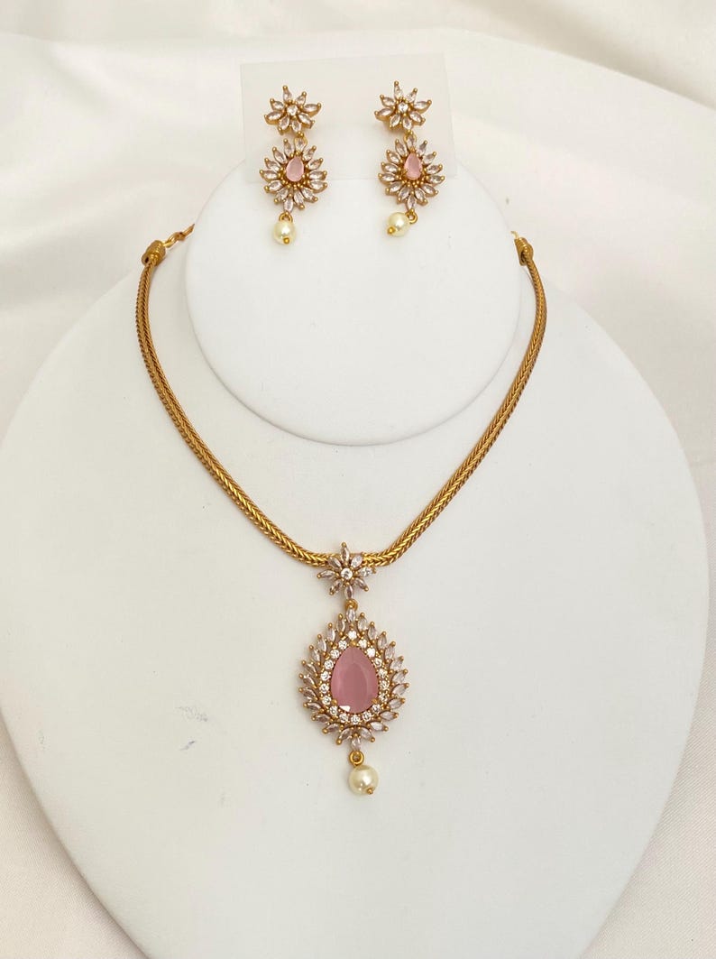May include: A gold-toned jewelry set featuring a necklace and matching earrings. The necklace has a chain and a teardrop pendant with a pink center stone surrounded by clear crystals. The earrings have a floral design with pink stones and dangling pearls.