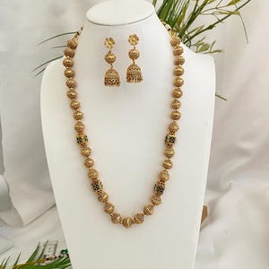 May include: Gold-toned beaded necklace and matching earrings set. The necklace features large, textured beads with small, decorative accents. The earrings have a floral top and a bell-shaped bottom. The jewelry is displayed on a white bust.