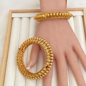 May include: Two gold-colored bangles with intricate floral and beaded designs. One bangle is open, while the other is closed and worn on a wrist. The jewelry is displayed on a light-colored surface, showcasing the detailed craftsmanship.