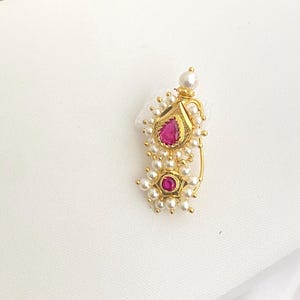 May include: A gold-colored nose ring featuring a teardrop-shaped design with a vibrant pink gemstone at its center. The ring is adorned with small, round, white pearls and a smaller pink gemstone at the bottom.