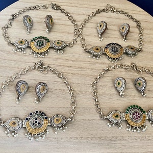 May include: Three silver necklaces with matching earrings. Each necklace features a circular pendant with a gold centre and green, blue, or pink accents. The earrings are teardrop shaped with a gold centre and green accents.