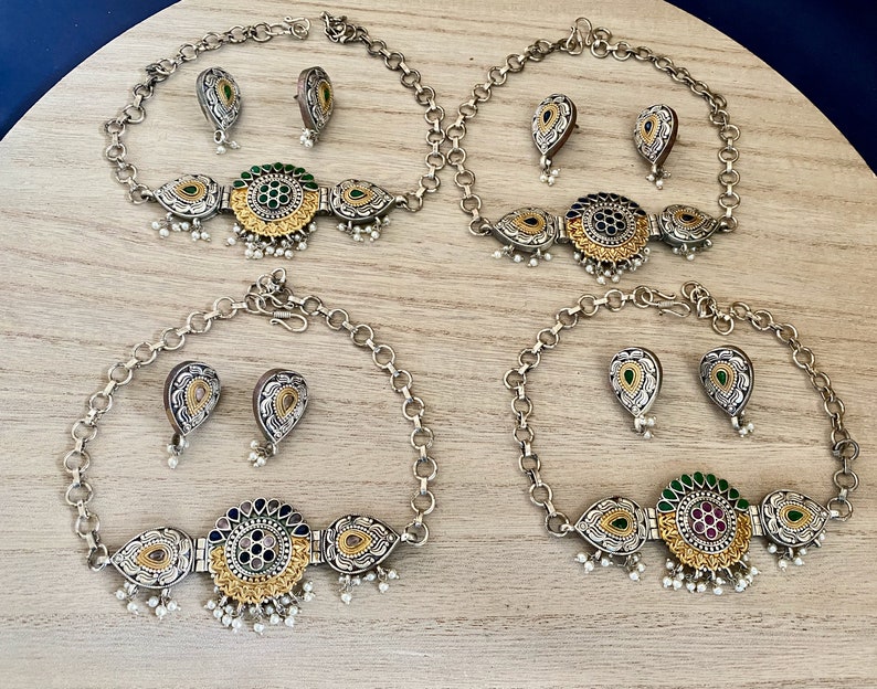 May include: Three silver necklaces with matching earrings. Each necklace features a circular pendant with a gold centre and green, blue, or pink accents. The earrings are teardrop shaped with a gold centre and green accents.