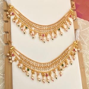 May include: Gold-toned ear chains with multiple strands, adorned with small pearls and colorful accents of red and green. The chains are displayed on a white surface, showcasing the intricate design and craftsmanship of the jewelry.