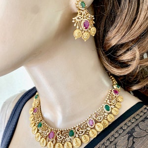 May include: Gold necklace and earrings set with red and green gemstones. The necklace features a design of coins and peacocks.