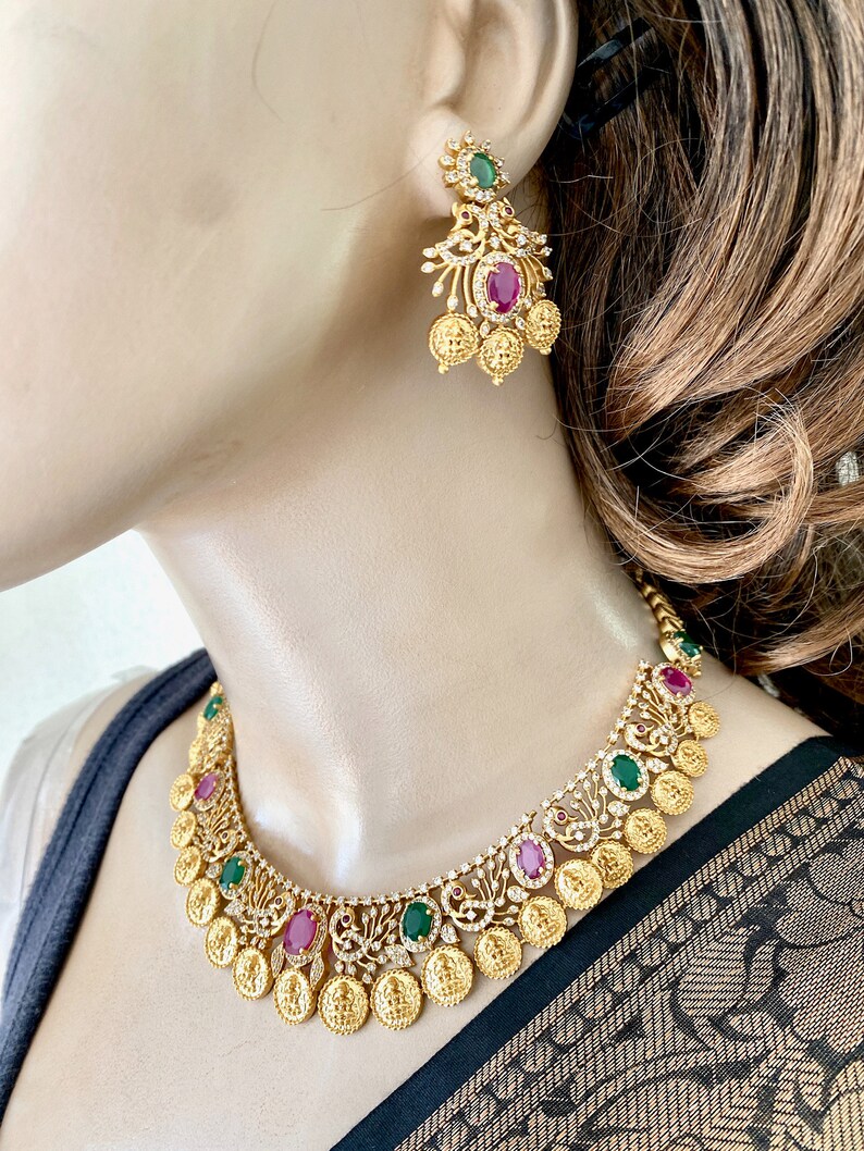 May include: Gold necklace and earrings set with red and green gemstones. The necklace features a design of coins and peacocks.