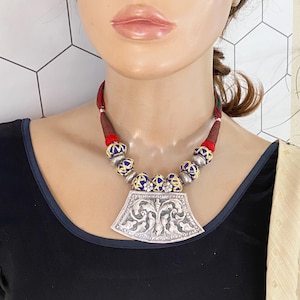 May include: A silver pendant necklace with a blue and gold patterned design. The pendant is shaped like a fan and has intricate floral details. The necklace is made with red and blue fabric cord and silver beads.
