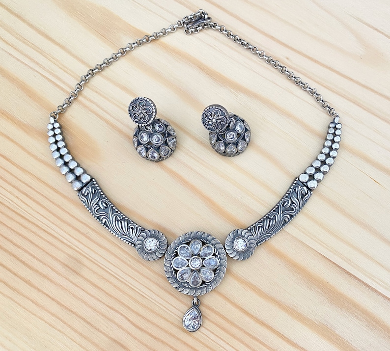 May include: A silver necklace with a floral design and a teardrop pendant. The necklace is adorned with clear stones and comes with matching earrings.