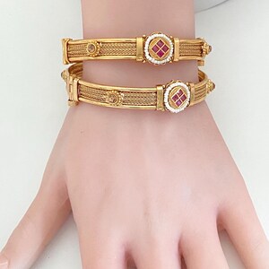 May include: Two gold-toned bangles featuring a woven metal texture, each centered with a circular medallion containing four small red square stones surrounded by tiny white beads. The bangles also include small round amber-colored accents.