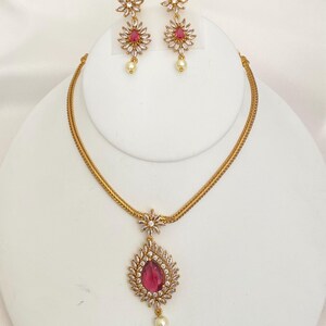 May include: A gold-toned jewelry set featuring a necklace and matching earrings. The necklace has a pendant with a teardrop-shaped, ruby-colored stone surrounded by clear crystals. The earrings and necklace have a floral design with a pearl drop.