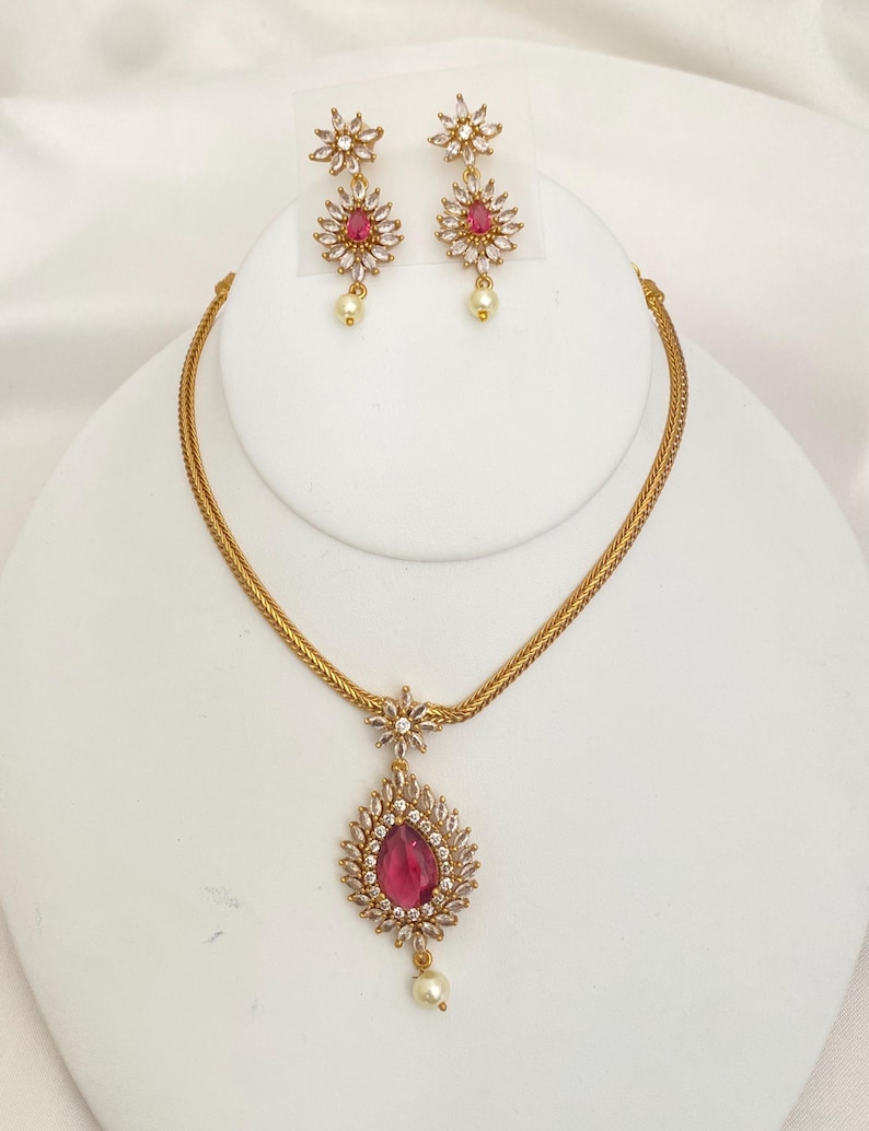 May include: A gold-toned jewelry set featuring a necklace and matching earrings. The necklace has a pendant with a teardrop-shaped, ruby-colored stone surrounded by clear crystals. The earrings and necklace have a floral design with a pearl drop.