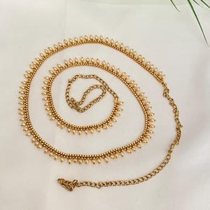 May include: Gold-toned body chain with small, pearl-like beads and a chain closure. The chain is arranged in a spiral on a white surface, with green leaves in the background. The chain is designed to be worn around the waist.