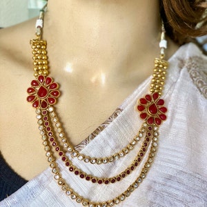 Puede incluir: Un collar de oro de varias capas con detalles de flores rojas y piedras transparentes. El collar est&aacute; sobre un fondo de tela blanca.