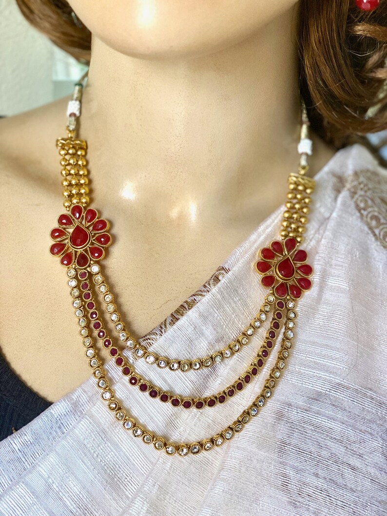 Puede incluir: Un collar de oro de varias capas con detalles de flores rojas y piedras transparentes. El collar est&aacute; sobre un fondo de tela blanca.