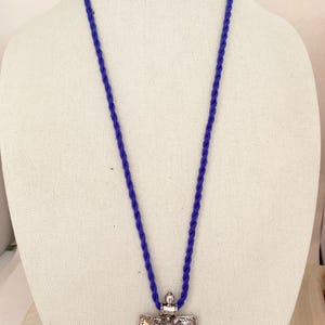May include: A silver pendant necklace with an ornate design hangs from a blue cord. The pendant is shaped like a shield and has intricate details.