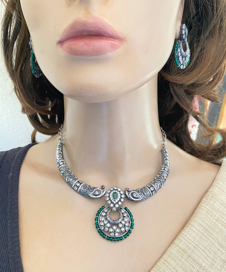 May include: Silver necklace with a green and white pendant. The pendant is a circle with a smaller circle inside. The necklace is on a mannequin.
