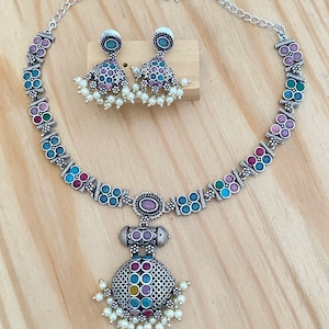 May include: A silver necklace with a round pendant featuring colorful stones and a pearl drop. Matching earrings with colorful stones and pearl drops are also included.