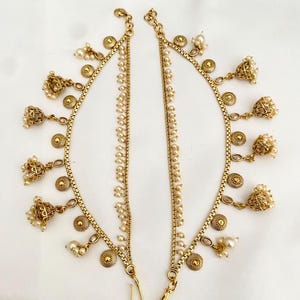 May include: A pair of gold-colored anklets with intricate detailing. Each anklet features a chain with small, round, pearl-like beads and decorative elements, including small bells and circular accents. The anklets are designed to be worn around the ankle.