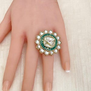 May include: A gold ring with a large, clear, square-shaped stone set in a green enamel frame. The stone is surrounded by a ring of smaller, white stones.