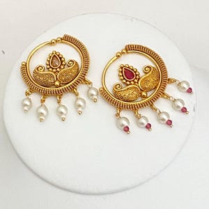 May include: A pair of gold-toned earrings with a circular design. Each earring features a central teardrop-shaped stone, ornate detailing, and dangling pearls and small red beads. The earrings are displayed on a white surface.