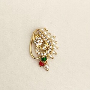 May include: A gold-tone nose ring featuring a curved paisley-shaped frame encrusted with clear crystals. The design is accented with a row of small white pearls along the outer edge and includes three dangling charms in green, red, and white.