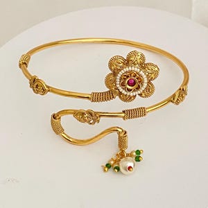 May include: A gold-tone open bangle bracelet featuring a central flower motif with a small red stone and white bead accents. The bracelet includes textured wire detailing and a dangling charm with a large white pearl and small green beads.