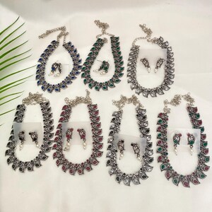 以下が含まれることがあります： シルバーカラーのネックレスとイヤリングのコレクション。各ネックレスは、青、緑、赤、クリアストーンなどのさまざまな色のアクセントが付いた涙の形をしたデザインが特徴です。イヤリングも同様のデザインで、小さなパールのアクセントが付いています。