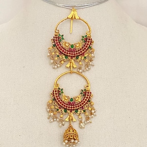May include: Gold-toned earrings with a crescent-shaped design. The earrings feature red and green accents, small white beads, and a bell-shaped charm. The earrings are displayed on a white surface.