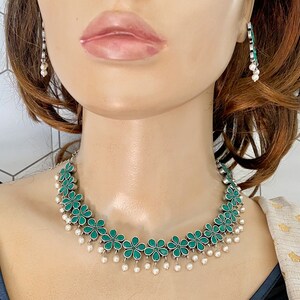 Op de afbeelding: Een zilveren ketting met groene bloemvormige hangers en witte parel druppels. De ketting wordt gedragen door een model met bruin haar.