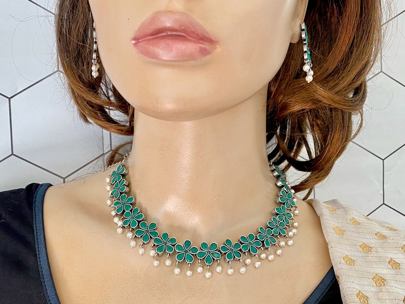 Op de afbeelding: Een zilveren ketting met groene bloemvormige hangers en witte parel druppels. De ketting wordt gedragen door een model met bruin haar.